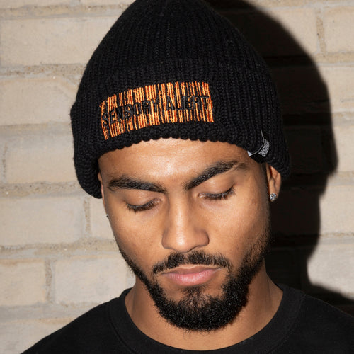 Man wearing a black knit beanie with a logo against a brick wall.